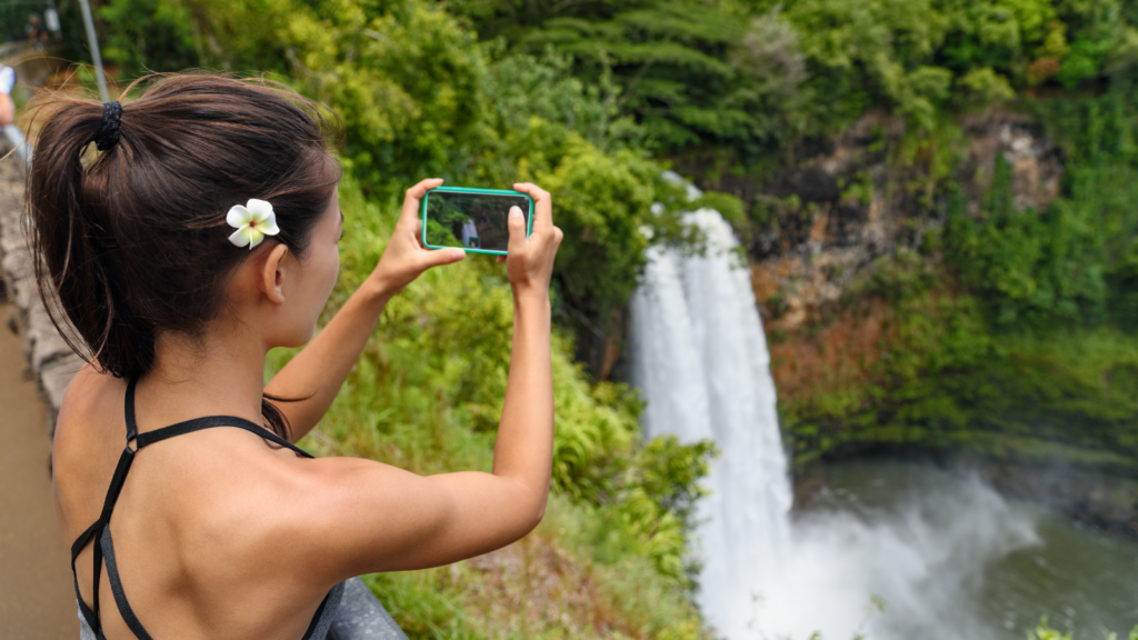 Women in Hawaii with flower in her hair taking a photo of a waterfall.