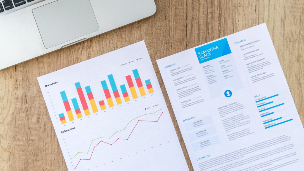 Table with resume printed in blue next to laptop and document with graph on it.