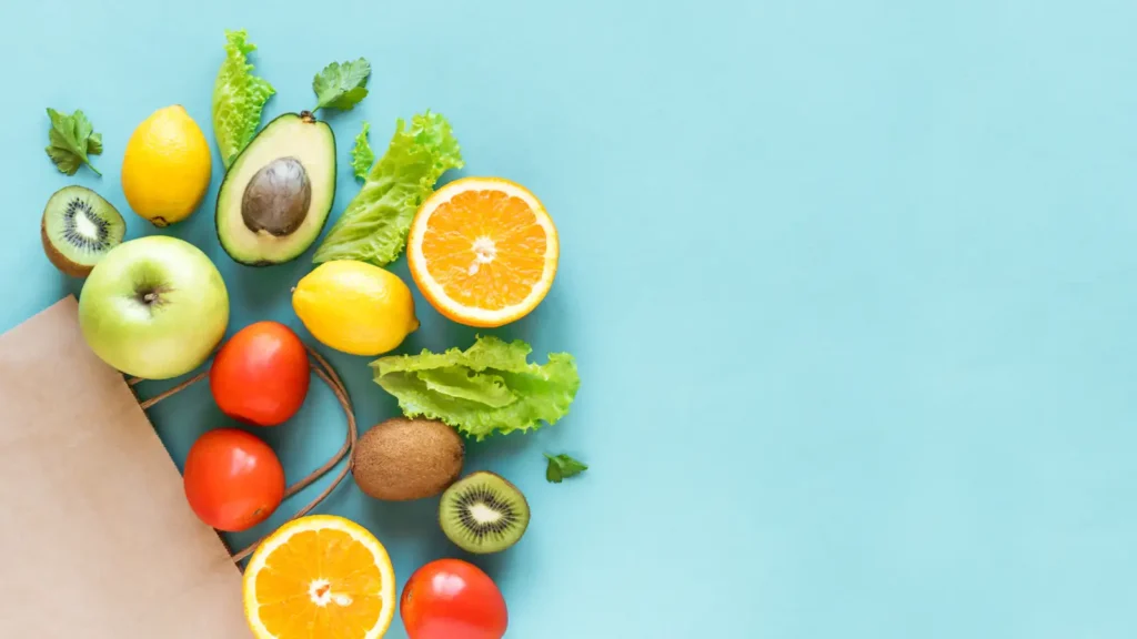 A variety of fruits and vegetables spilling out of a brown paper bag on a turquoise background.