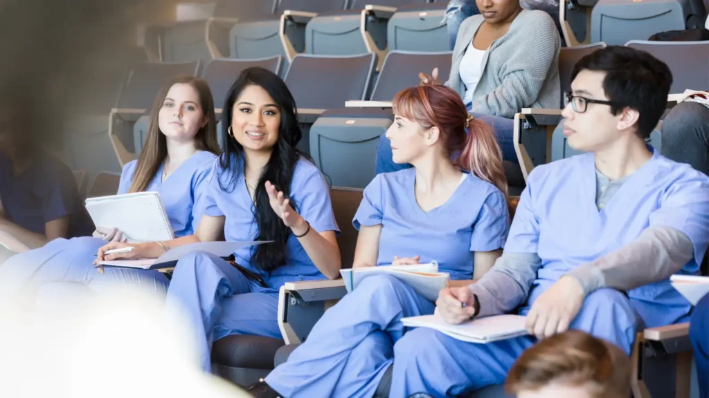 Travel nurses in blue medical scrubs sitting in a classroom with a travel nurse reaching out to ask a common question.